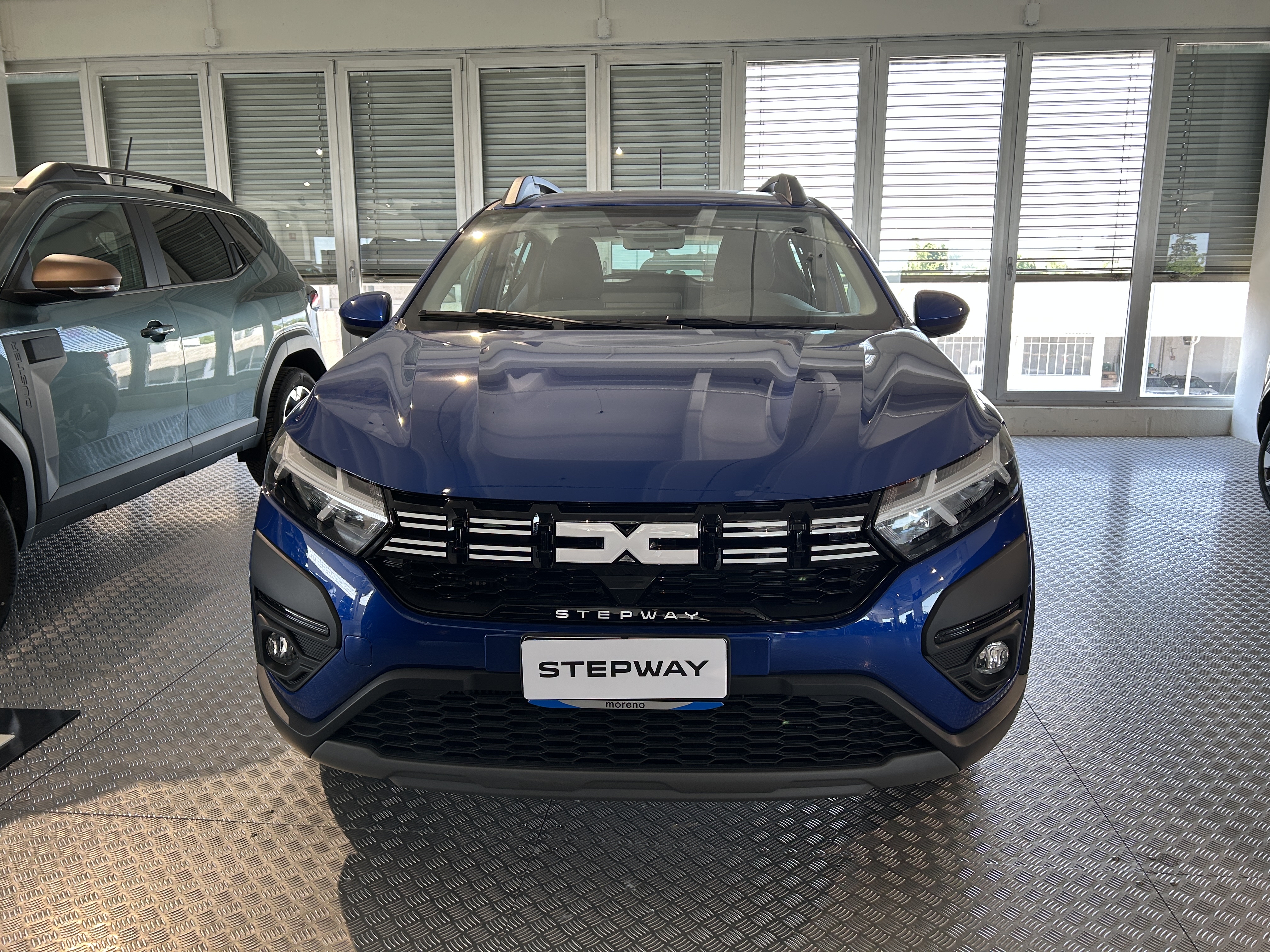 Vista frontale della nuova Dacia Sandero Stepway di colore blu esposta in showroom, con logo Dacia rinnovato sul frontale e design moderno.