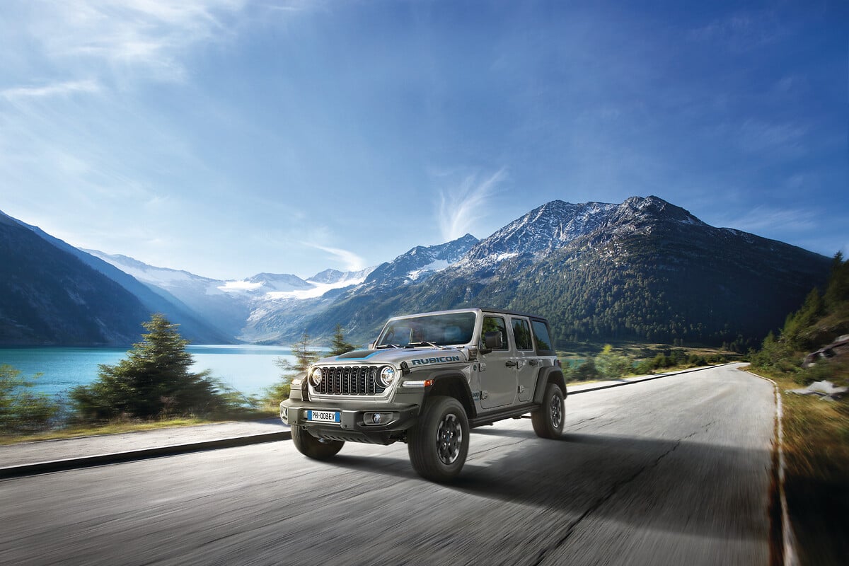 Jeep Wrangler Rubicon grigia su una strada di montagna con vista su un lago e alte vette innevate.