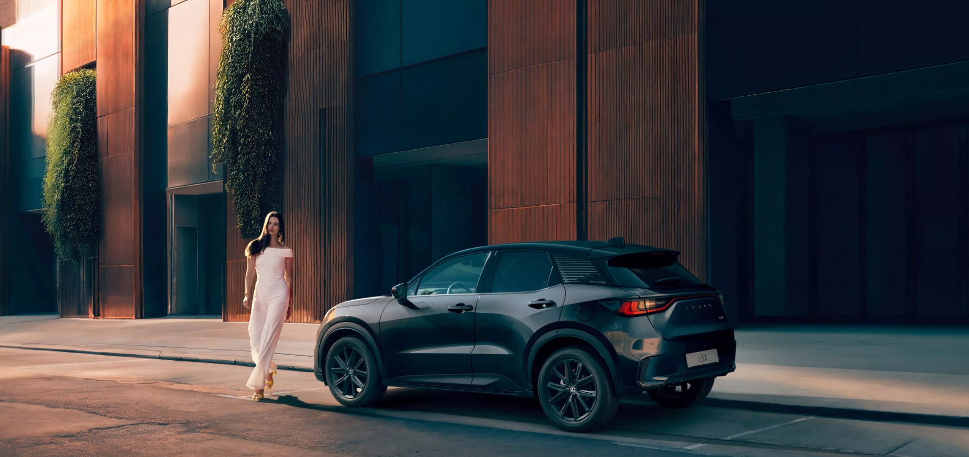 Lexus LBX Vibrant Edition vista posteriore laterale in un contesto urbano moderno, con una persona che cammina accanto all’auto. 