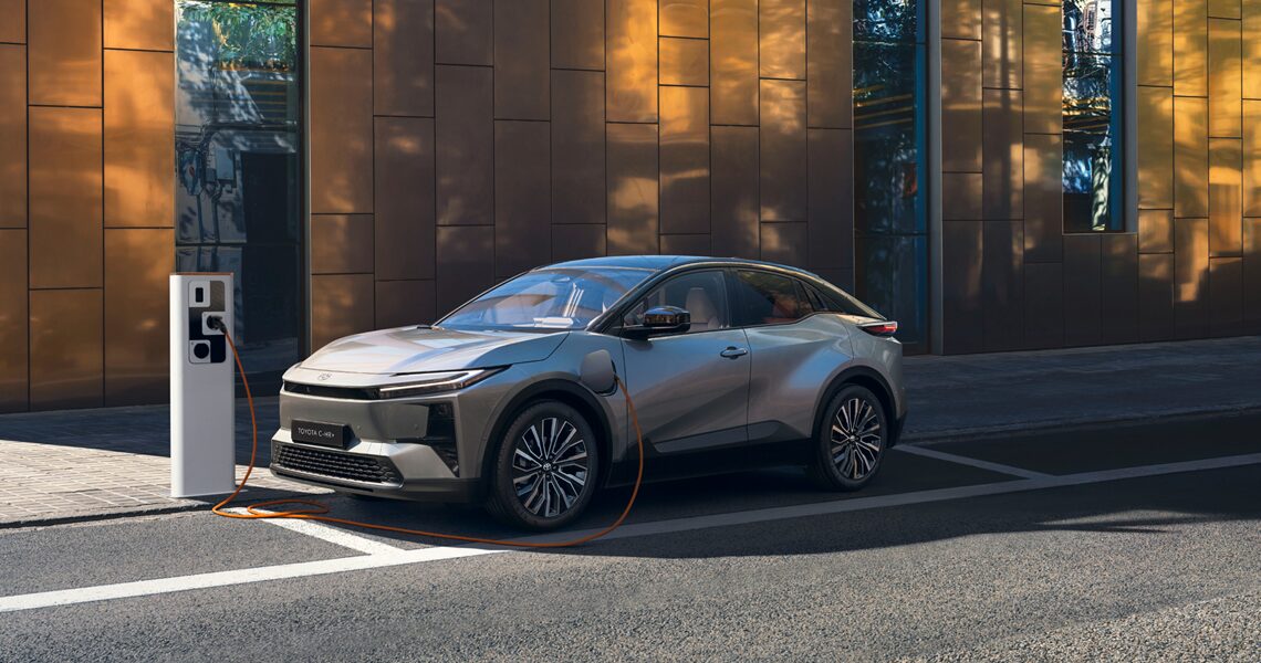 Toyota C-HR+ grigia in fase di ricarica su una colonnina elettrica davanti a un palazzo dallo stile contemporaneo 